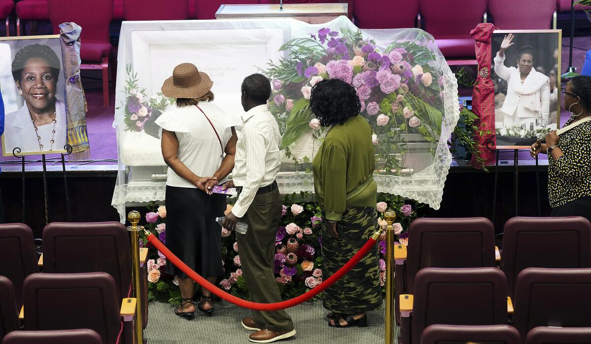 Three people stand before a white casket flanked by portraits of a woman and adorned with flowers 