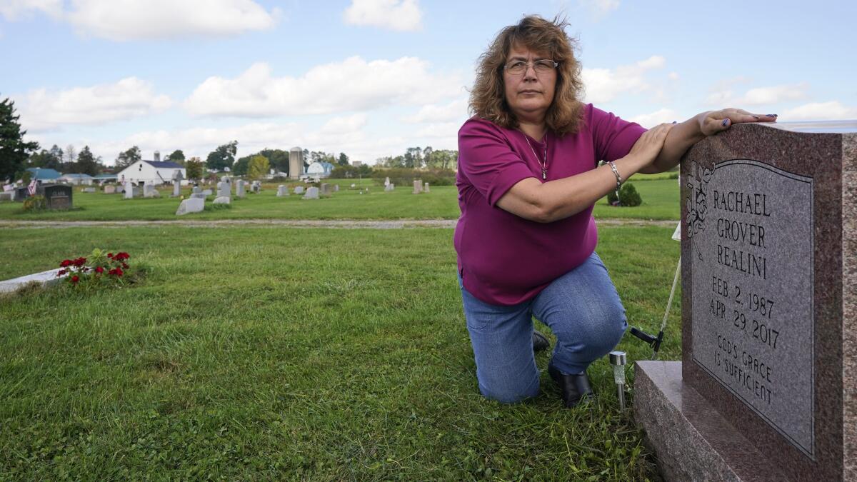 A woman kneels and places her hands on a cemetery gravestone. 