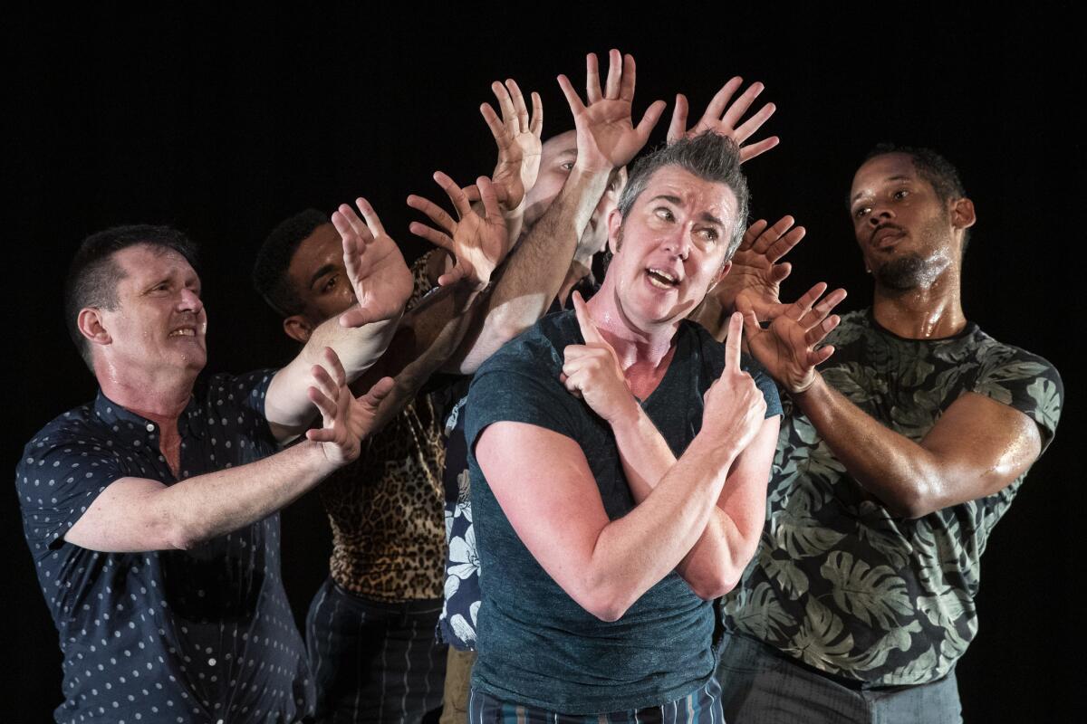 Sean Dorsey, center, stands, arms crossed and index fingers pointing up, with a group of men behind him, stretching their hands toward him.