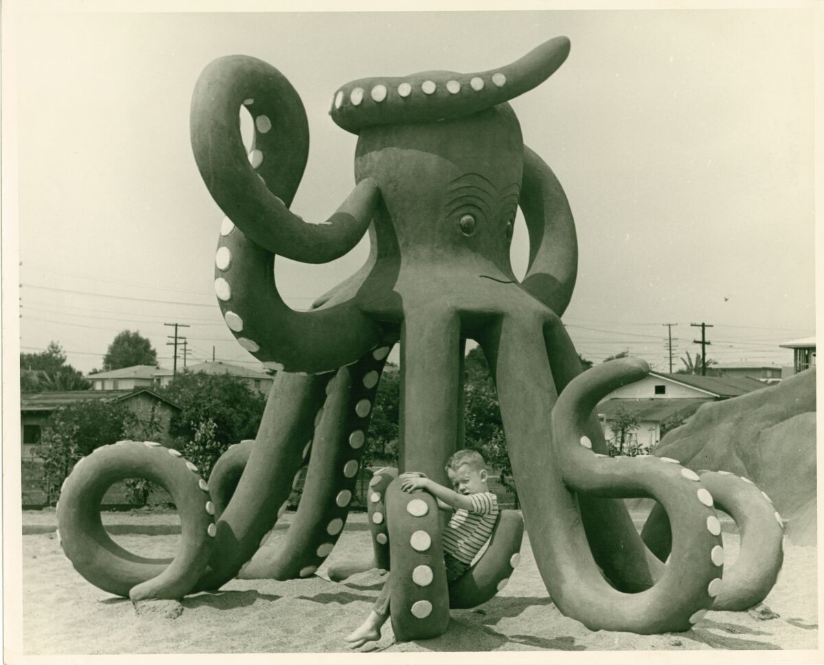 A child sits on the leg of a large sculpture of an octopus in a sandy area of a playground.