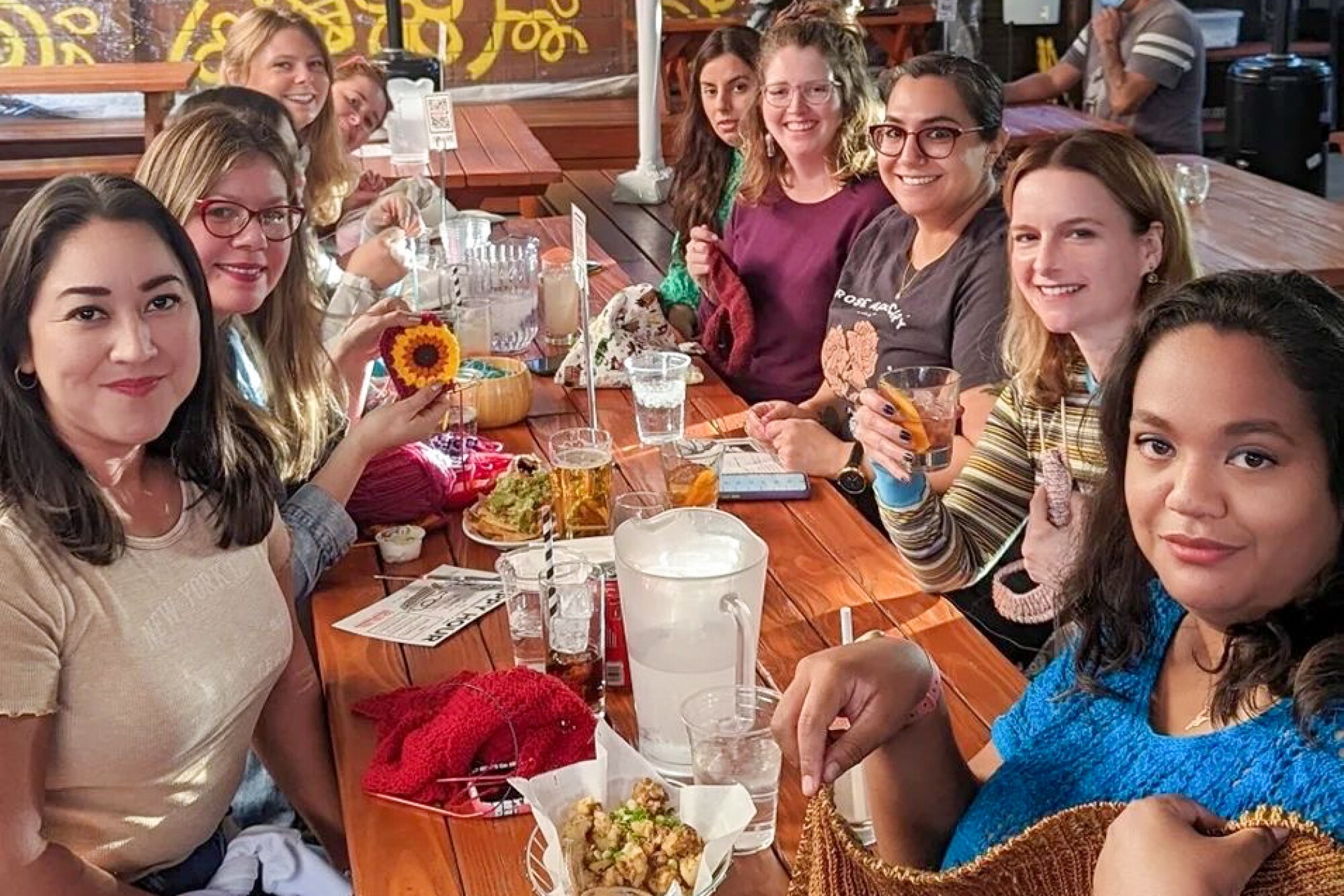 A group of people at a table with drinks and knitting projects.