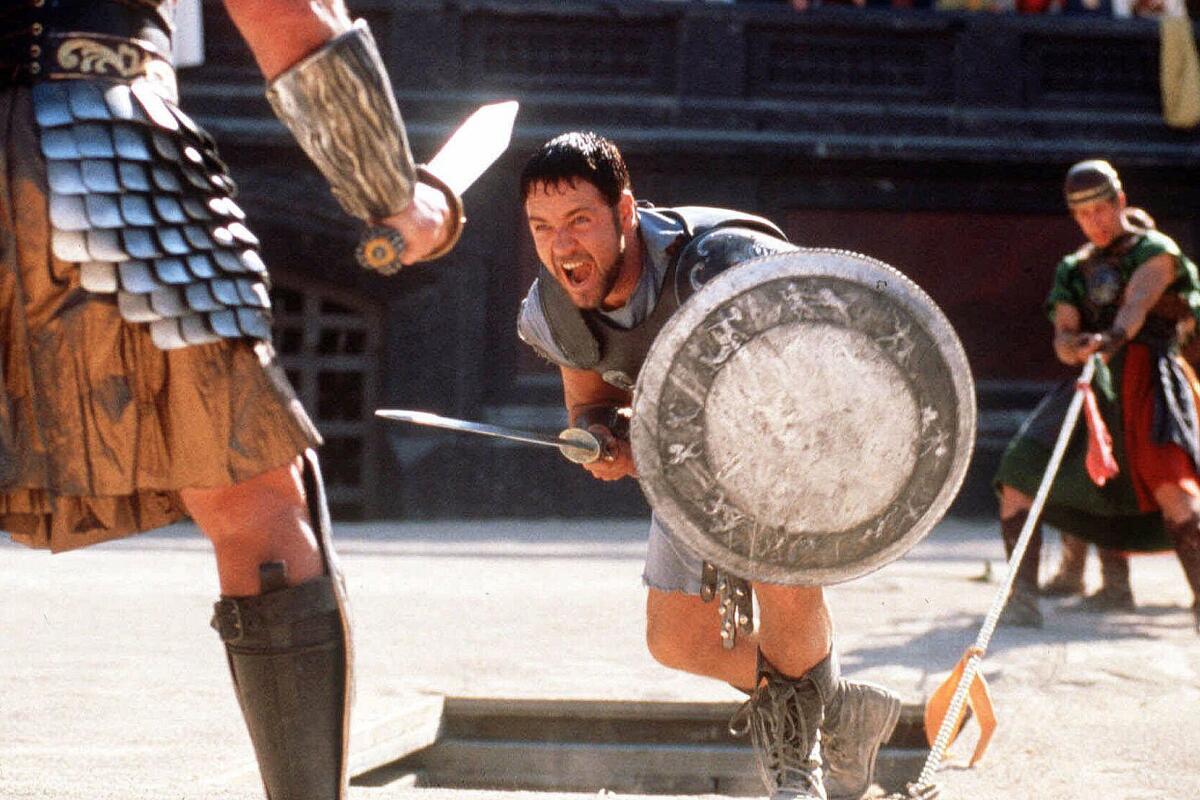 Russell Crowe, dressed as a gladiator, lunges forward with a sword and shield while yelling.