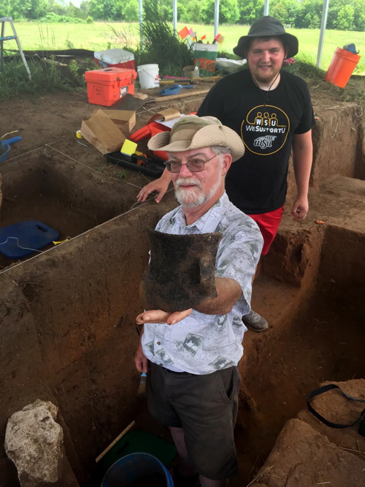 Professor Donald Blakeslee of Wichita State University shows a black pot unearthed by student Jeremiah Perkins, behind him.