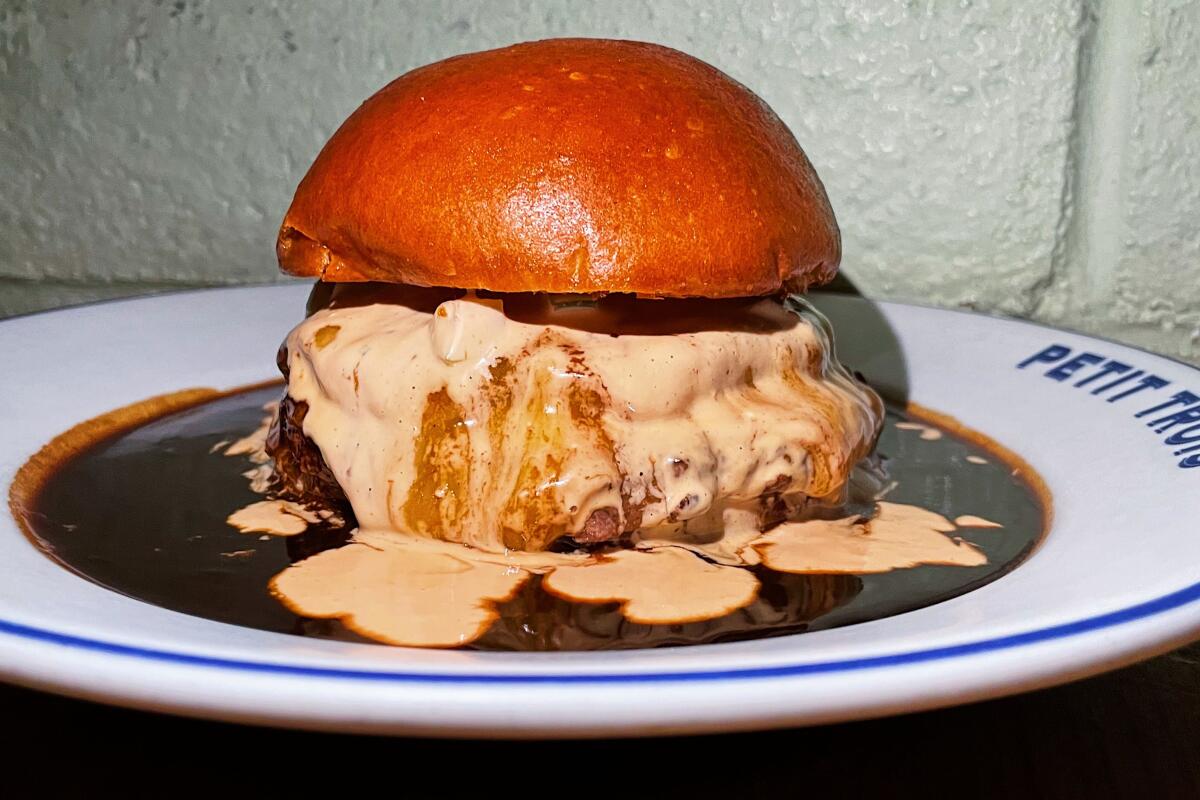 A horizontal closeup of a burger smothered in cream-colored and dark brown sauces beneath the bun on a "Petit Trois" plate