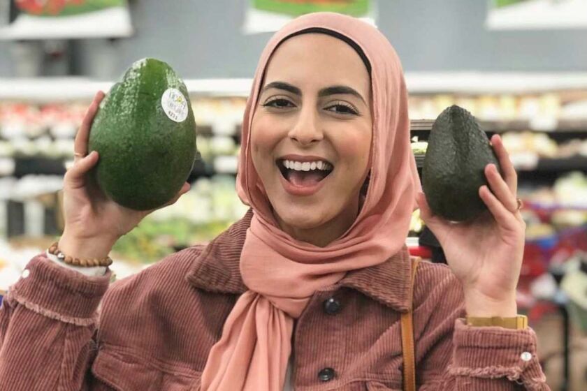 A woman in a hijab smiles in a grocery store as she holds a giant avocado in one hand and a regular-sized one in the other