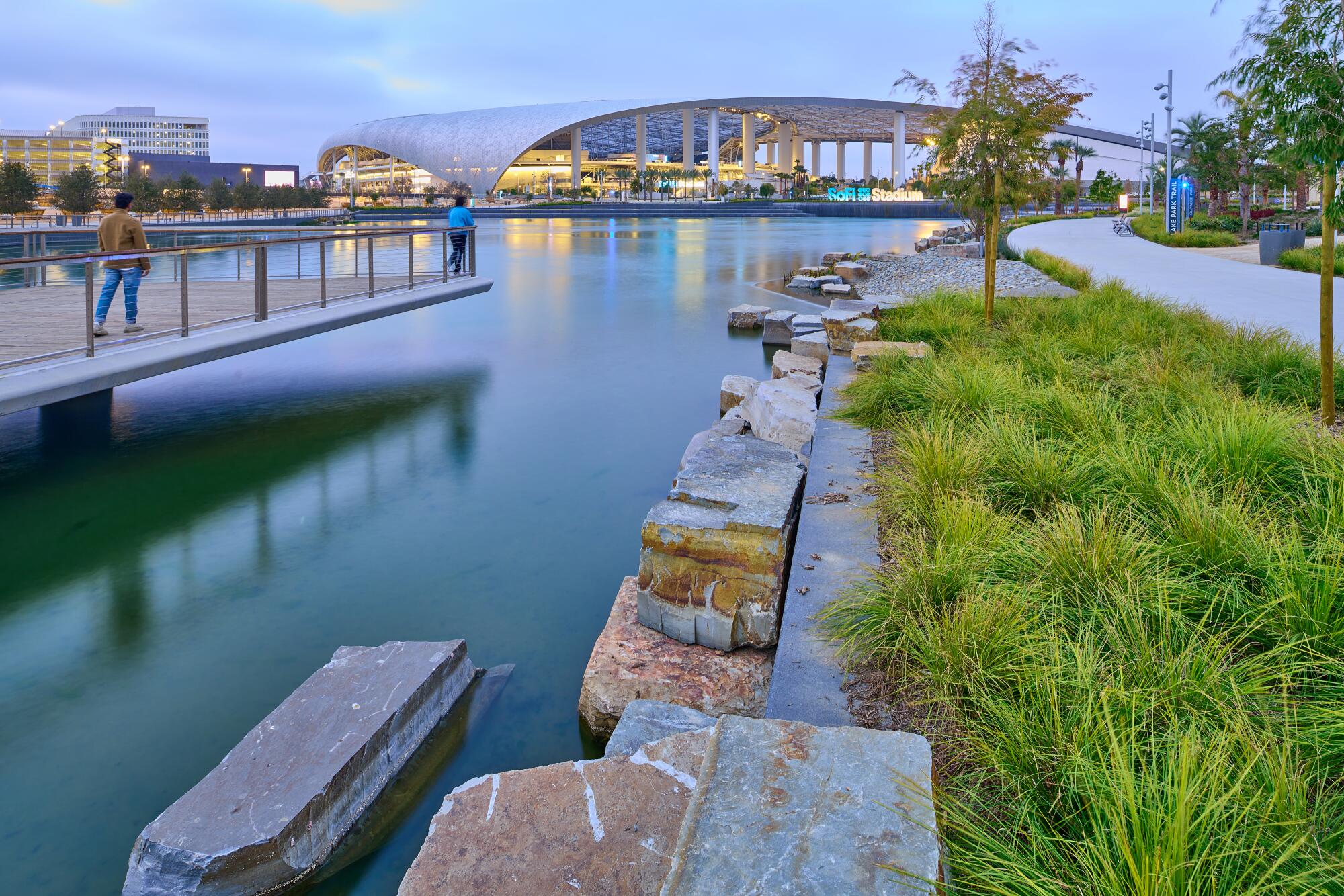 A lake surrounded by park sits before the curling form of SoFi Stadium's roof.