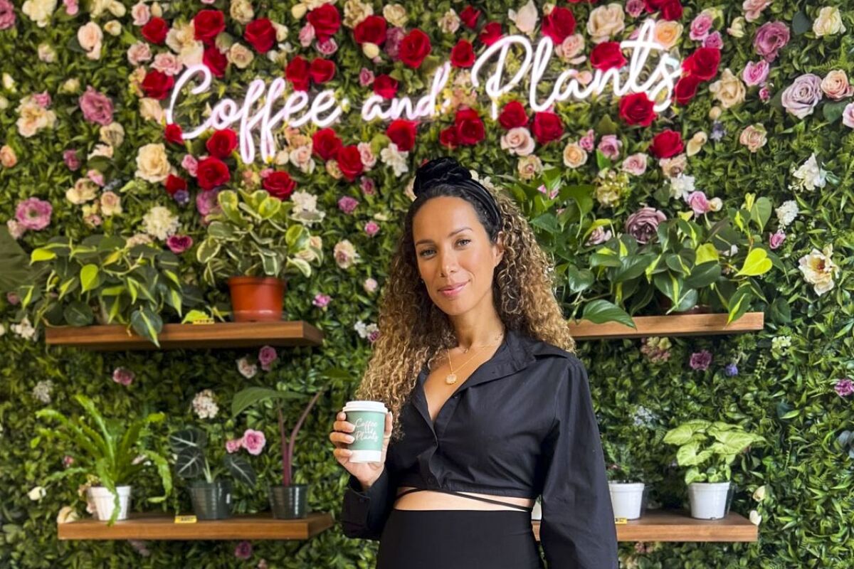 A smiling woman holds a cup in front of a plant wall with neon spelling out "Coffee and Plants"