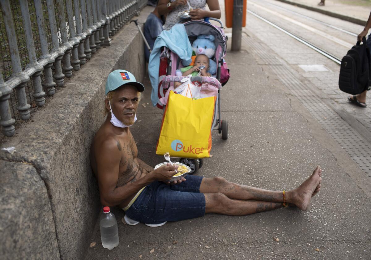 Man sits on sidewalk eating with face mask under his chin