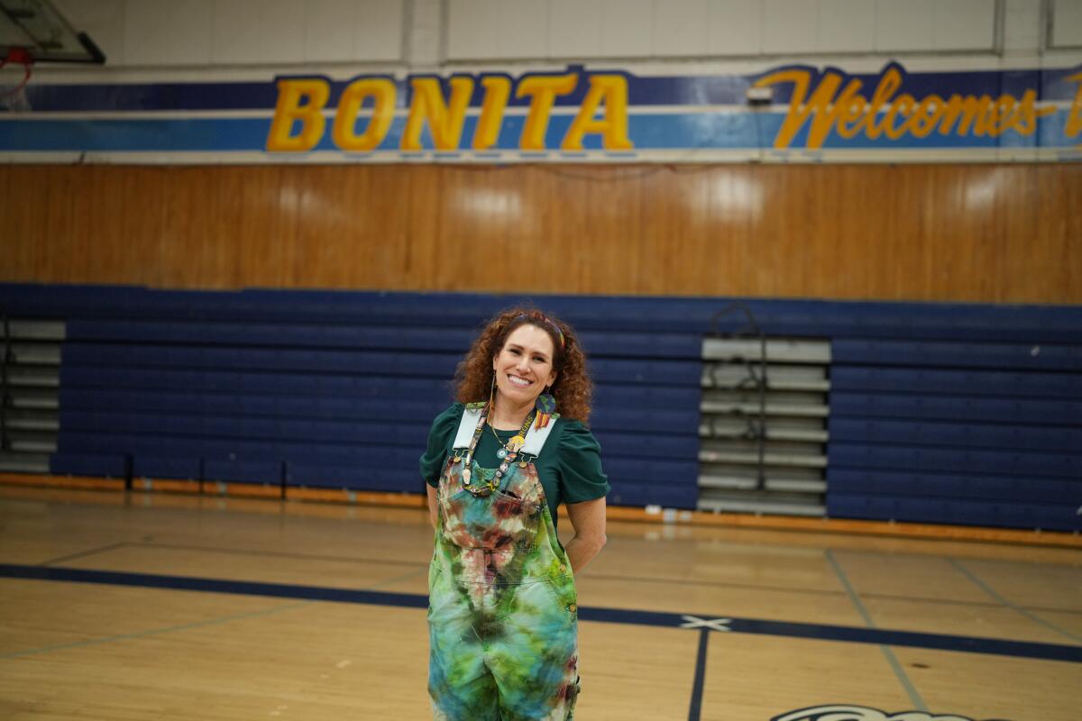 A woman wearing a tie-dyed smock stands in a school gym, smiling.