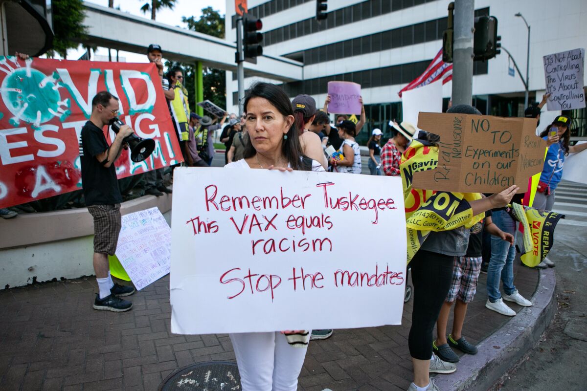 In L.A. a female protester holds a sign reading "Remember Tuskegee. This vax equals racism. Stop the mandates." 