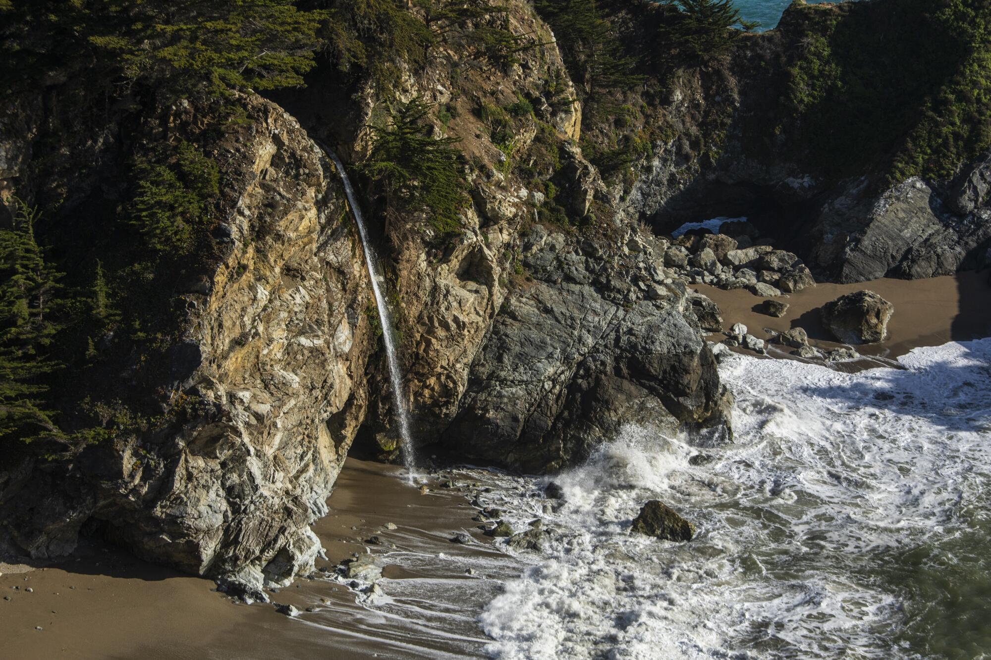 A narrow waterfall falls over rocks onto a beach.