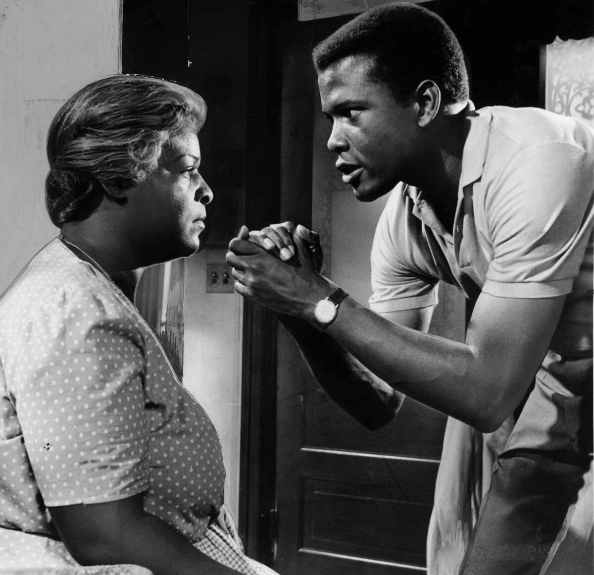 a black-and-white photo of a man standing up clasping his hands in front of a woman sitting down 