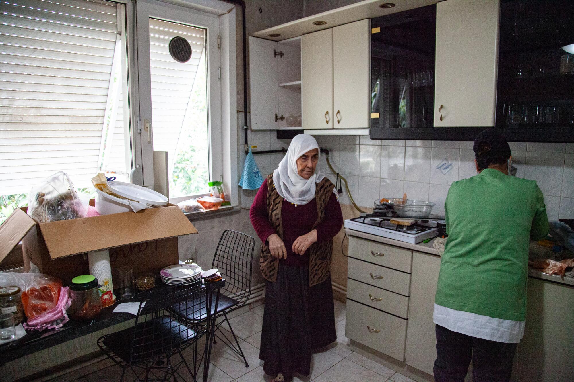 A woman in white headscarf stands in a kitchen with windows and white cabinets, with a box of belongings atop a table 