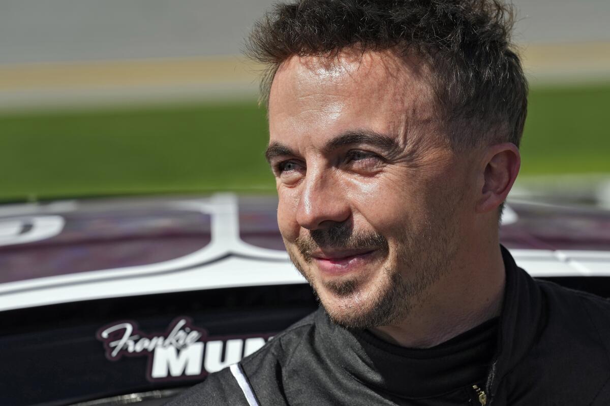 Frankie Muniz dressed in black smiles in the sun, standing in front of his race car at the Daytona Speedway