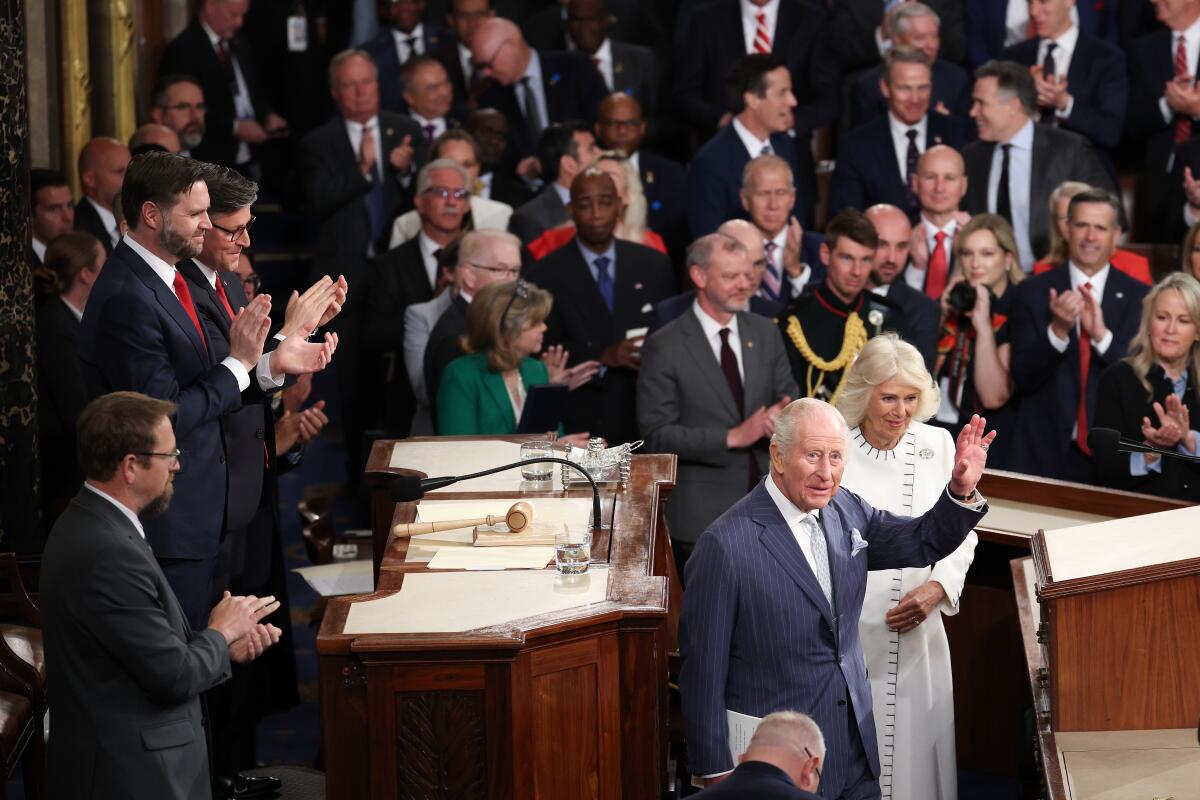 A man in a dark gray suit and pale blue tie waves next to a blond woman in a white dress, surrounded by people applauding