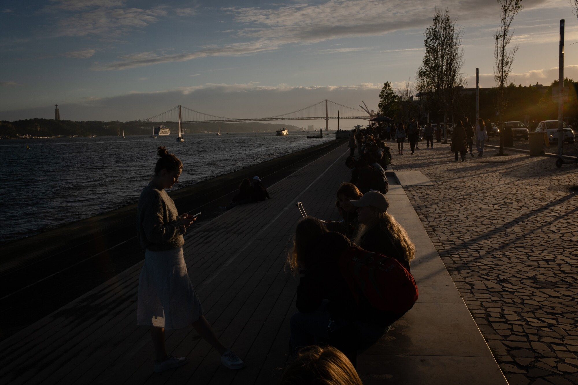 People congregate at dusk by a river, with views of a bridge 