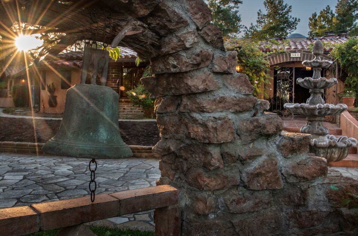 A bronze bell hangs near the entrance courtyard of an adobe home, with a four-tiered fountain in the background.