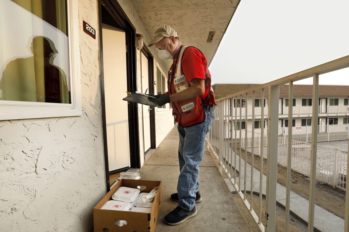 Red Cross volunteer wearing a mask and reflective vest stands outside a motel door with a clipboard and box of packaged meals