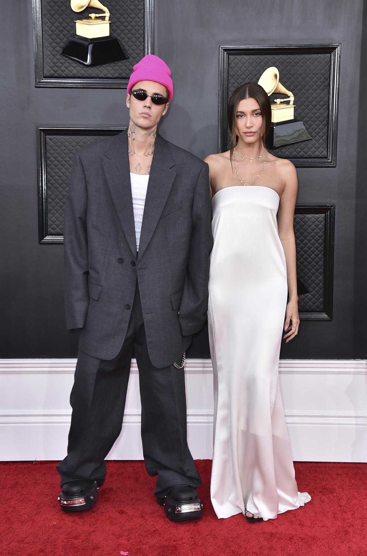 A man in an oversize suit and a pink beanie stands next to a woman in a white gown on a red carpet.