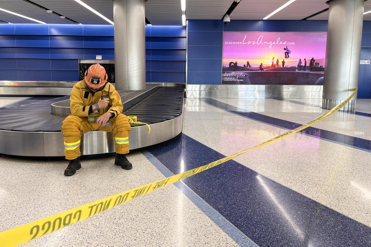 A firefighter sits on a still baggage conveyor behind yellow tape that reads, "Fire line do not cross."