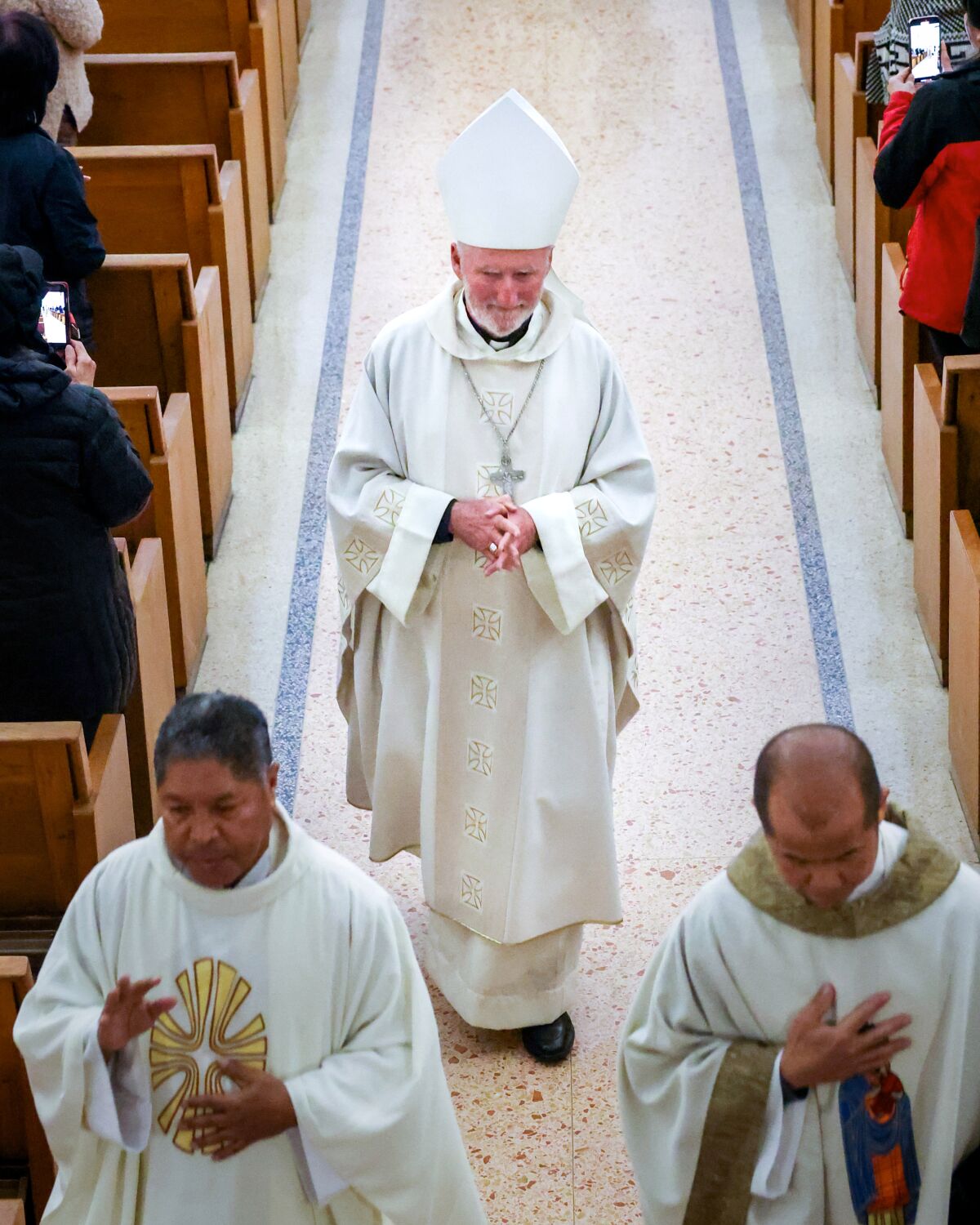 Three clergymen walk down the aisle of a church, between pews, toward the camera.