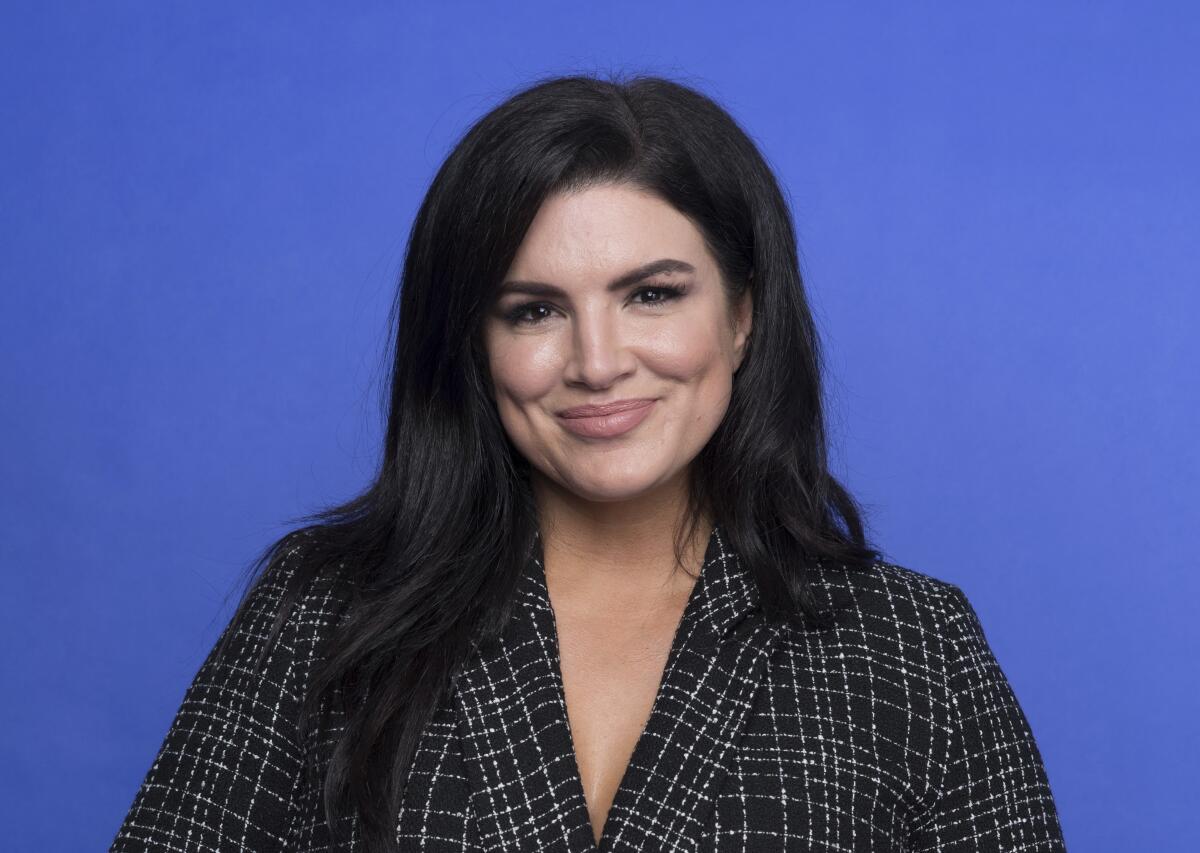 Gina Carano poses in a dark plaid suit jacket in front of a blue backdrop
