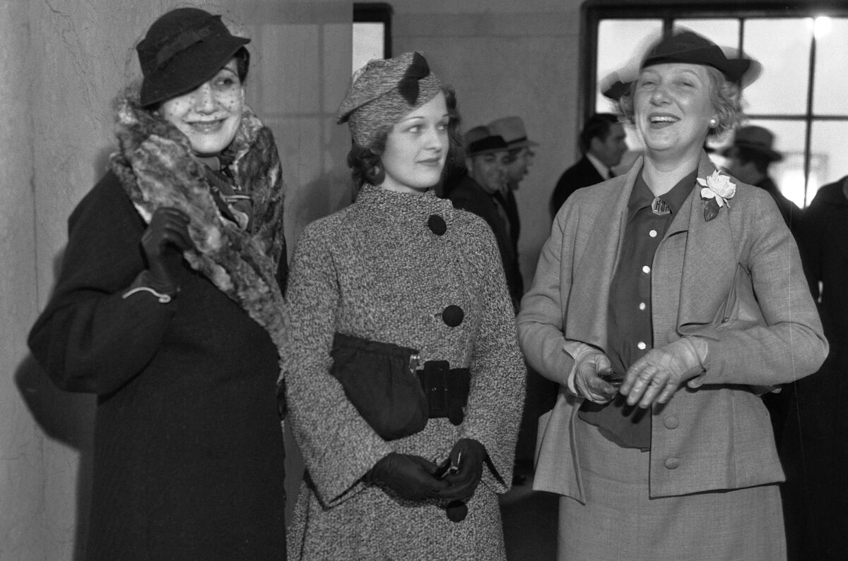 Three women in hats, gloves and one with a fur stole stand together, smiling and laughing, inside a court building.