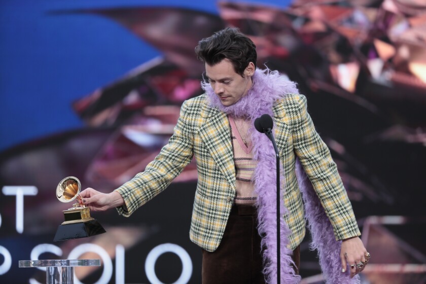 Harry Styles placing a Grammy on a podium while wearing a plaid blazer and lavender boa