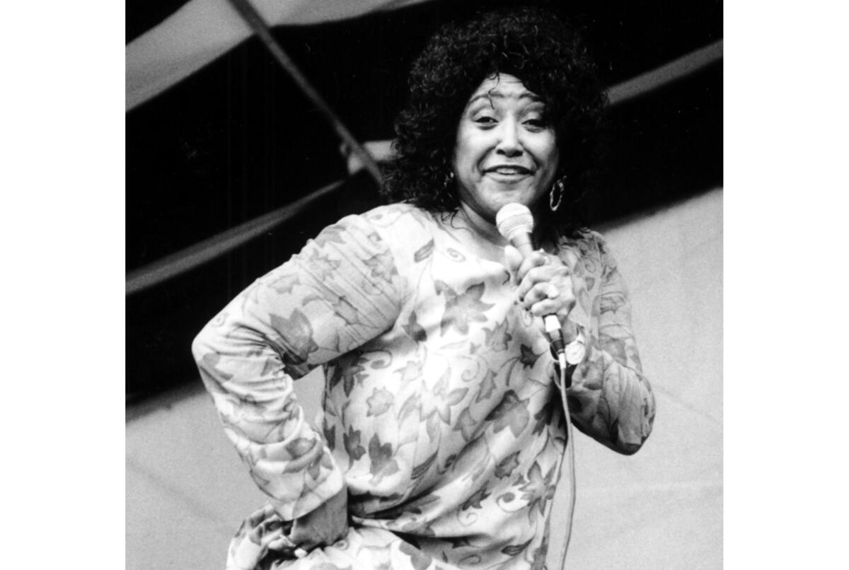 A black-and-white photo of a woman in a patterned dress with one hand on her hip singing into a hand-held microphone 