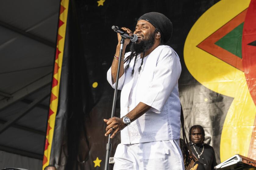 Peetah Morgan in a white ensemble and black cap holds a standing microphone while performing onstage.