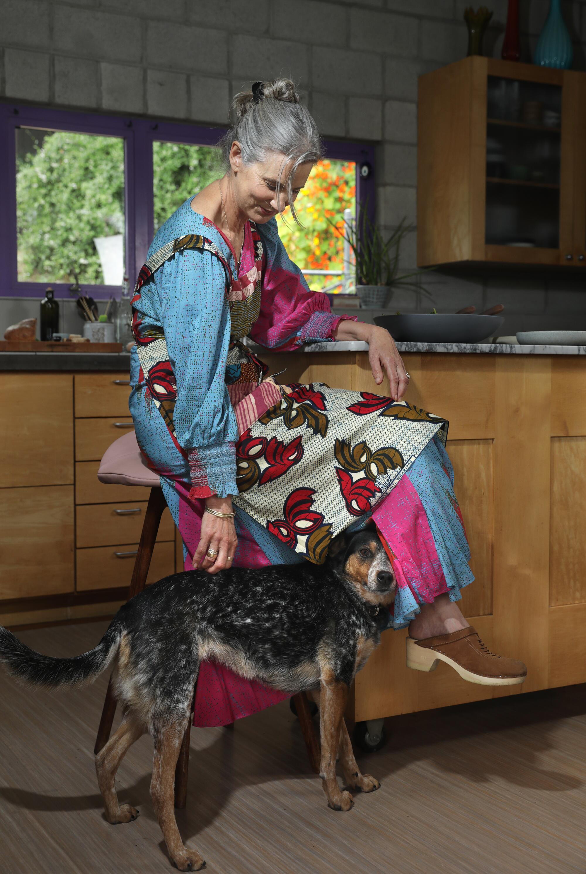 A woman seated at a kitchen counter bends down to pet her dog.