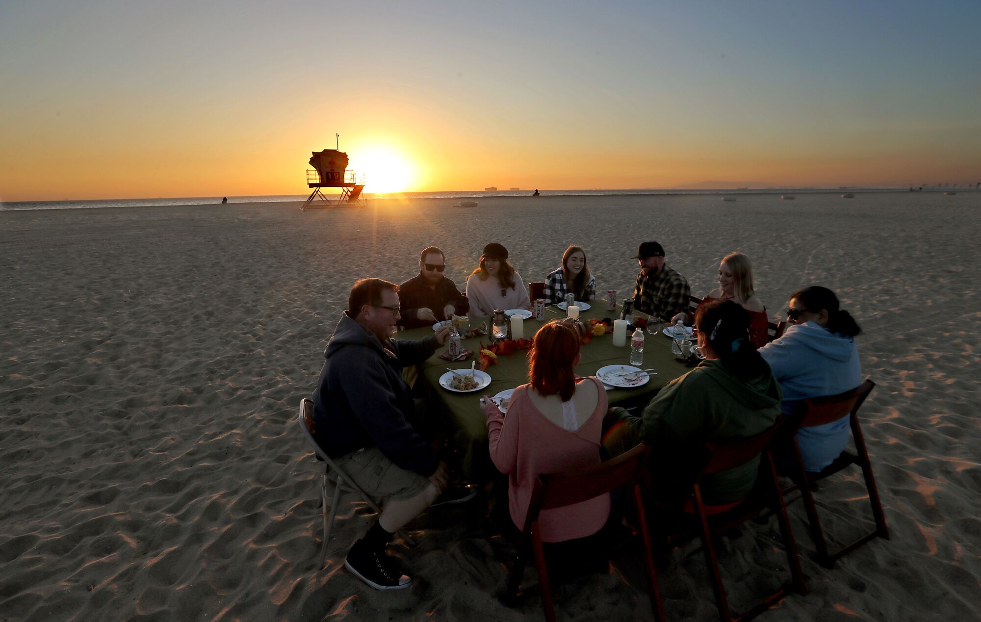 Nine people sit behind plates of food at a round table on the sand. In the background, the sun sets behind a lifeguard tower.