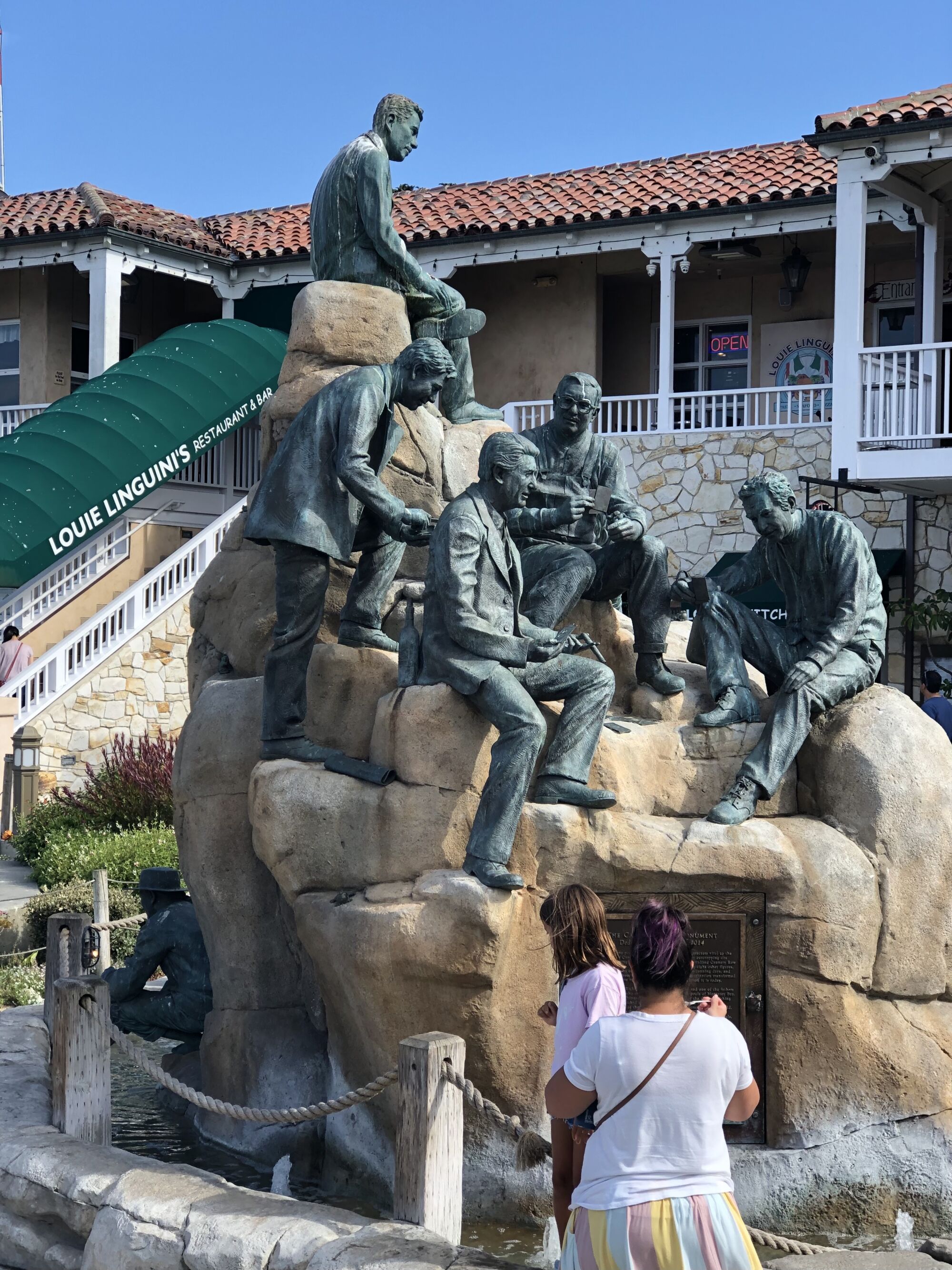 People passing a sculpture of several life-size figures on a large mound of tan boulders outside a restaurant.