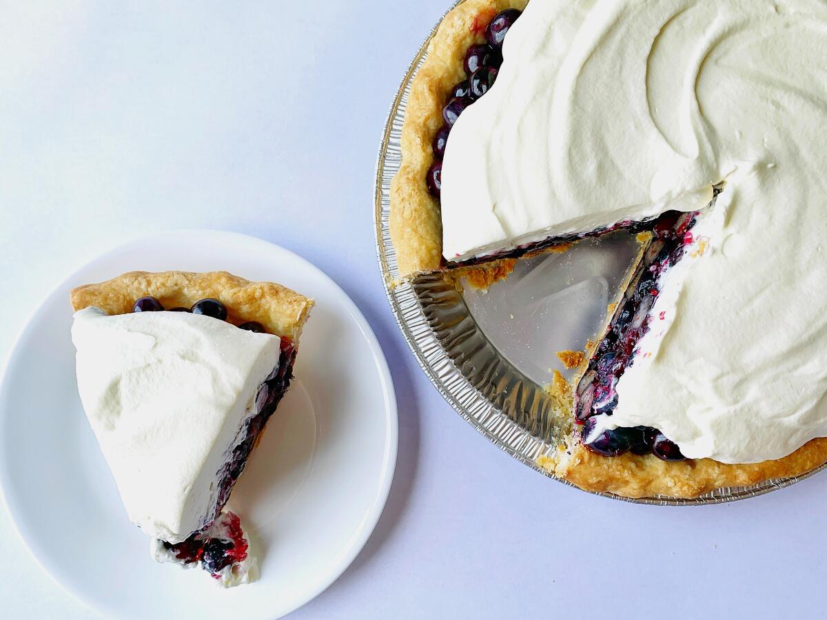 A slice of fresh blueberry pie with a creamy topping is served on a plate next to the remaining whole pie.