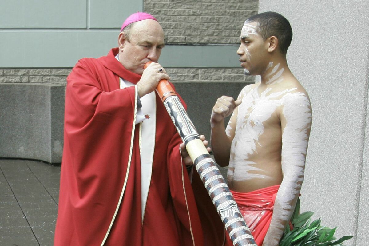 Bishop Christopher Alan Saunders tries to play a digeridoo while another man stands next to him.