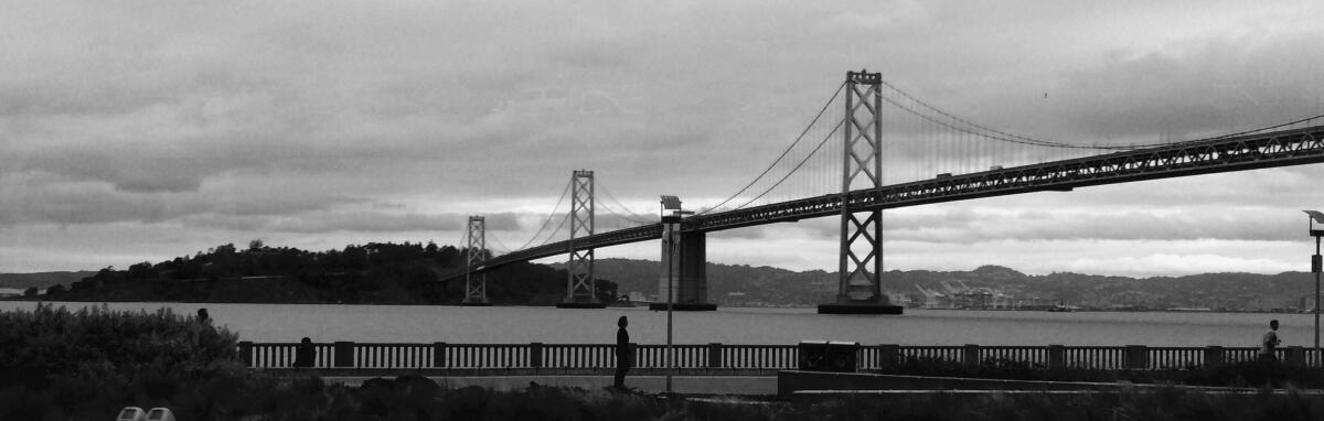 A large suspension bridge spans a section of the San Francisco Bay under cloudy skies.