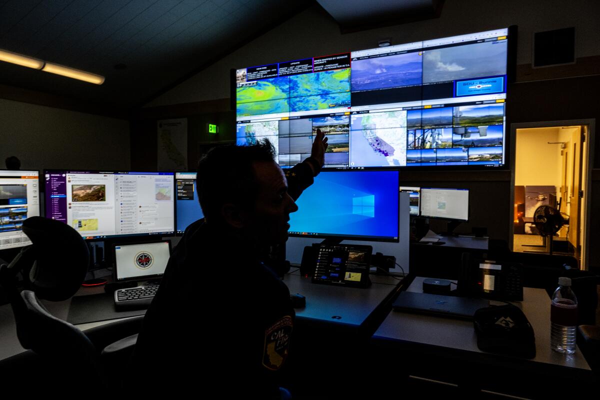 A man in a dimly light room turns away from his desk and gestures toward a wall of video monitors