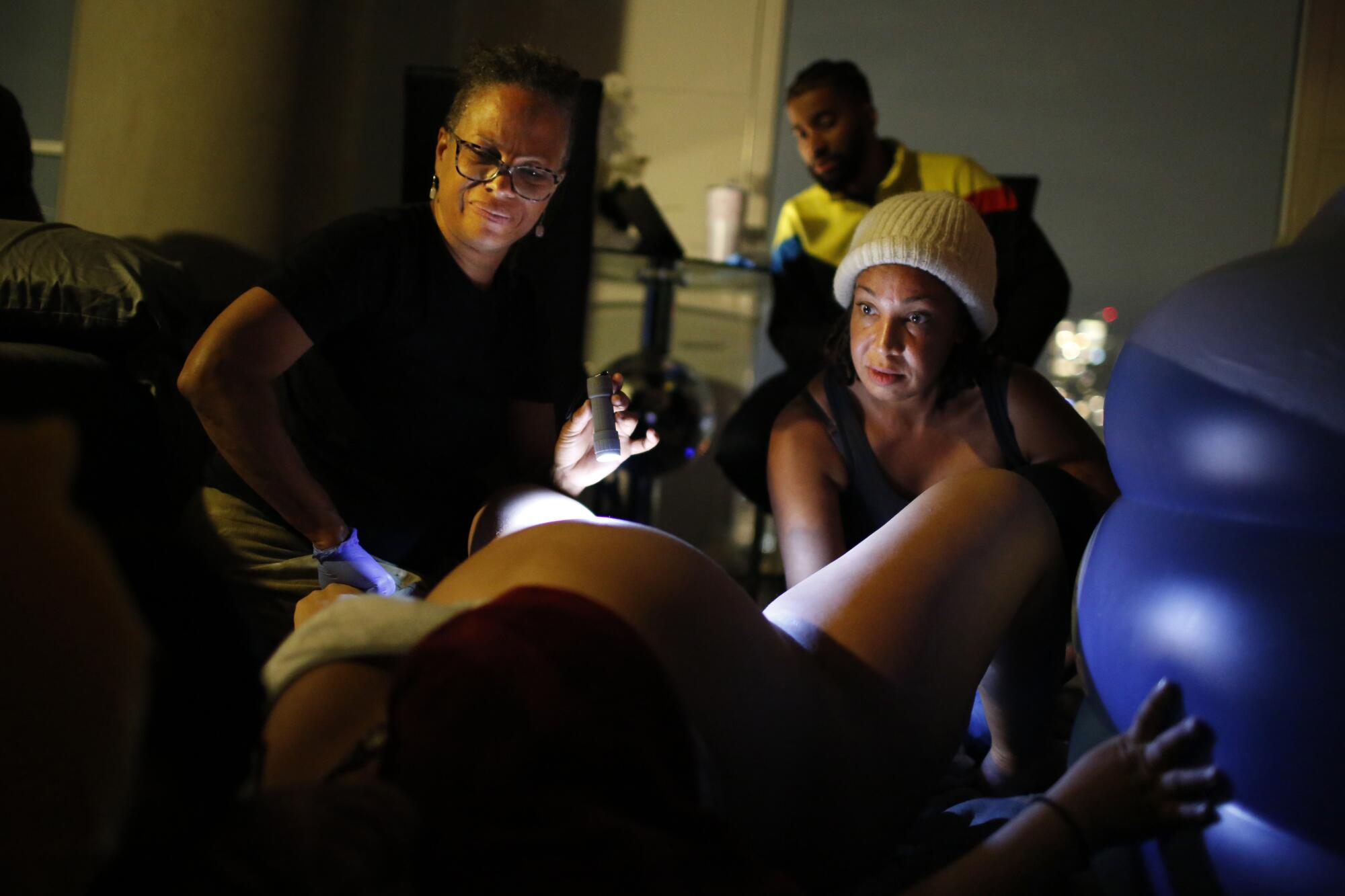 Two midwives, one holding a flashlight, assist a woman during labor