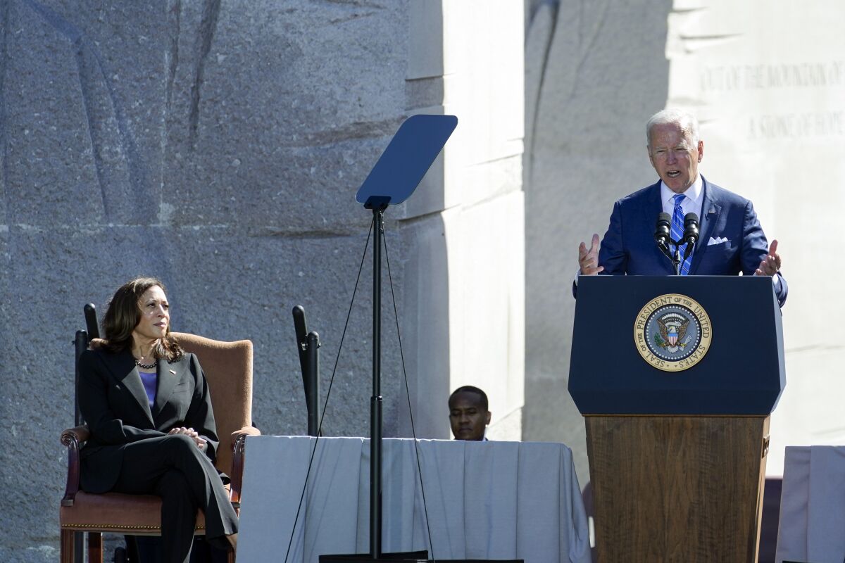 Vice President Kamala Harris sits and listens to President Biden speak at a podium.