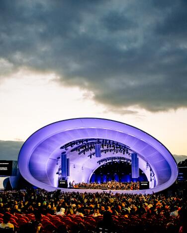 The Rady Shell at Jacobs Park lit a glowing blue, with rows of red chairs in front and the bay behind it