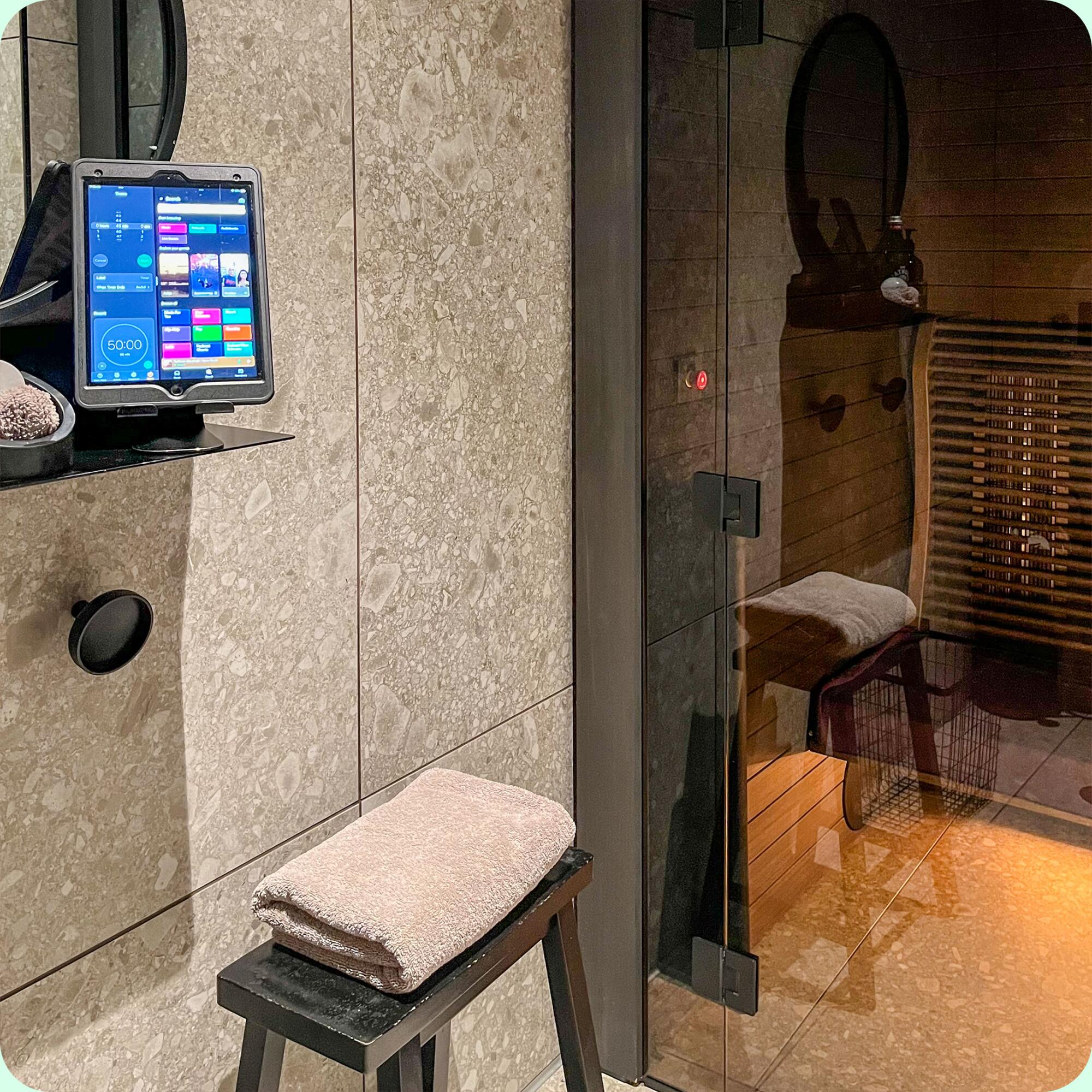 A glass door separates a sauna from a seat, mirror and iPad with a music player and timer.