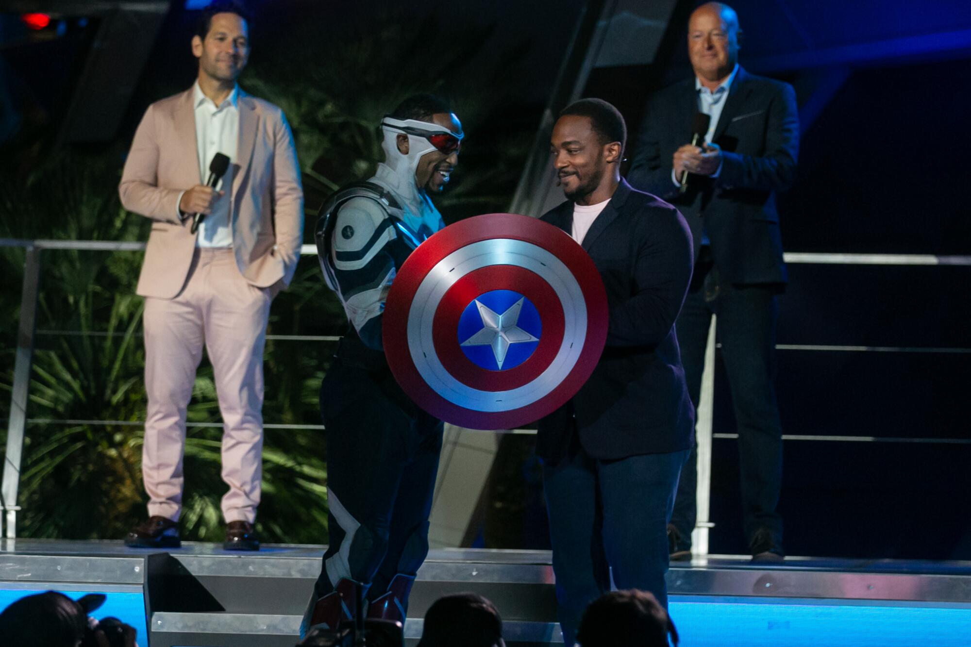 A smiling Anthony Mackie hands a Captain America shield to a Disney actor as Paul Rudd and a Disney executive look on.