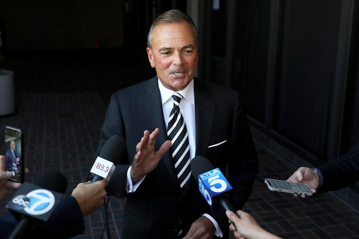 A man in a suit and tie faces a line of hands holding microphones with TV station logos on them.