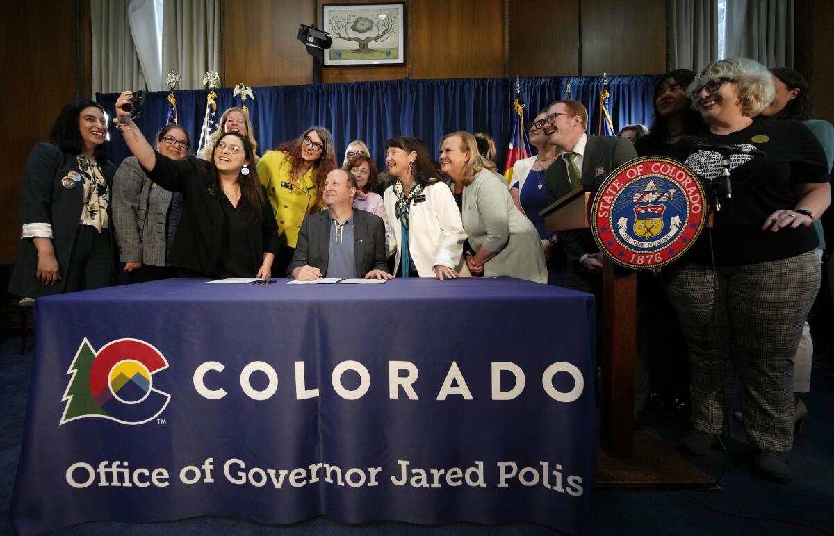 Governor Jared Polis, seated at a table, poses for a photo with others, standing 