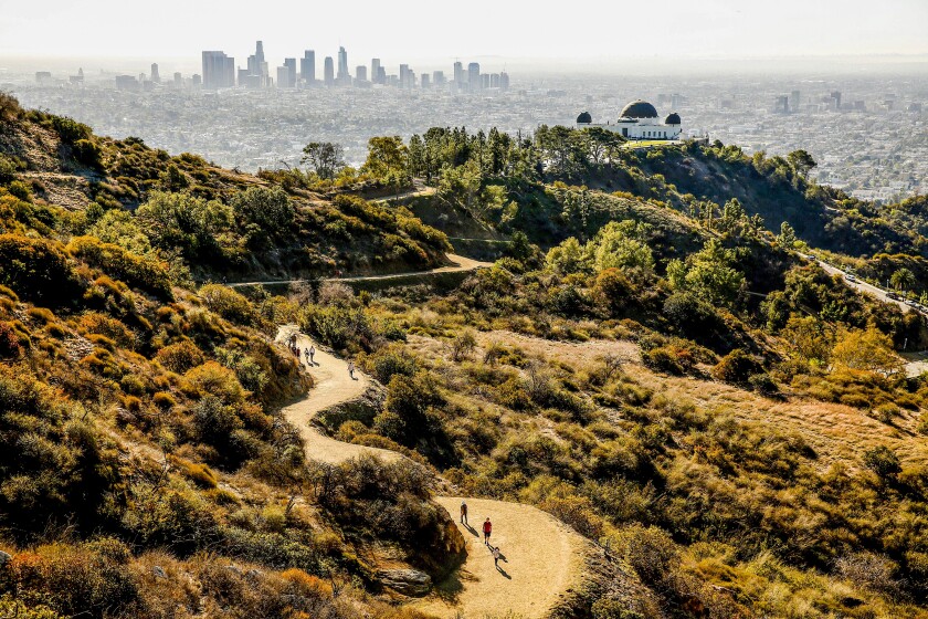 People hike on a trail in Griffith Park, with the observatory and the downtown skyline in the background.