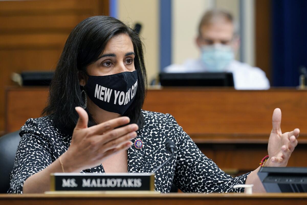 A woman speaks, seated at a desk, wearing a black face mask with the words "New York City."