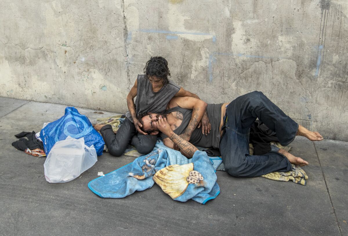 A man lies with his head in a woman's lap on a sidewalk surrounded by bags of their belongings