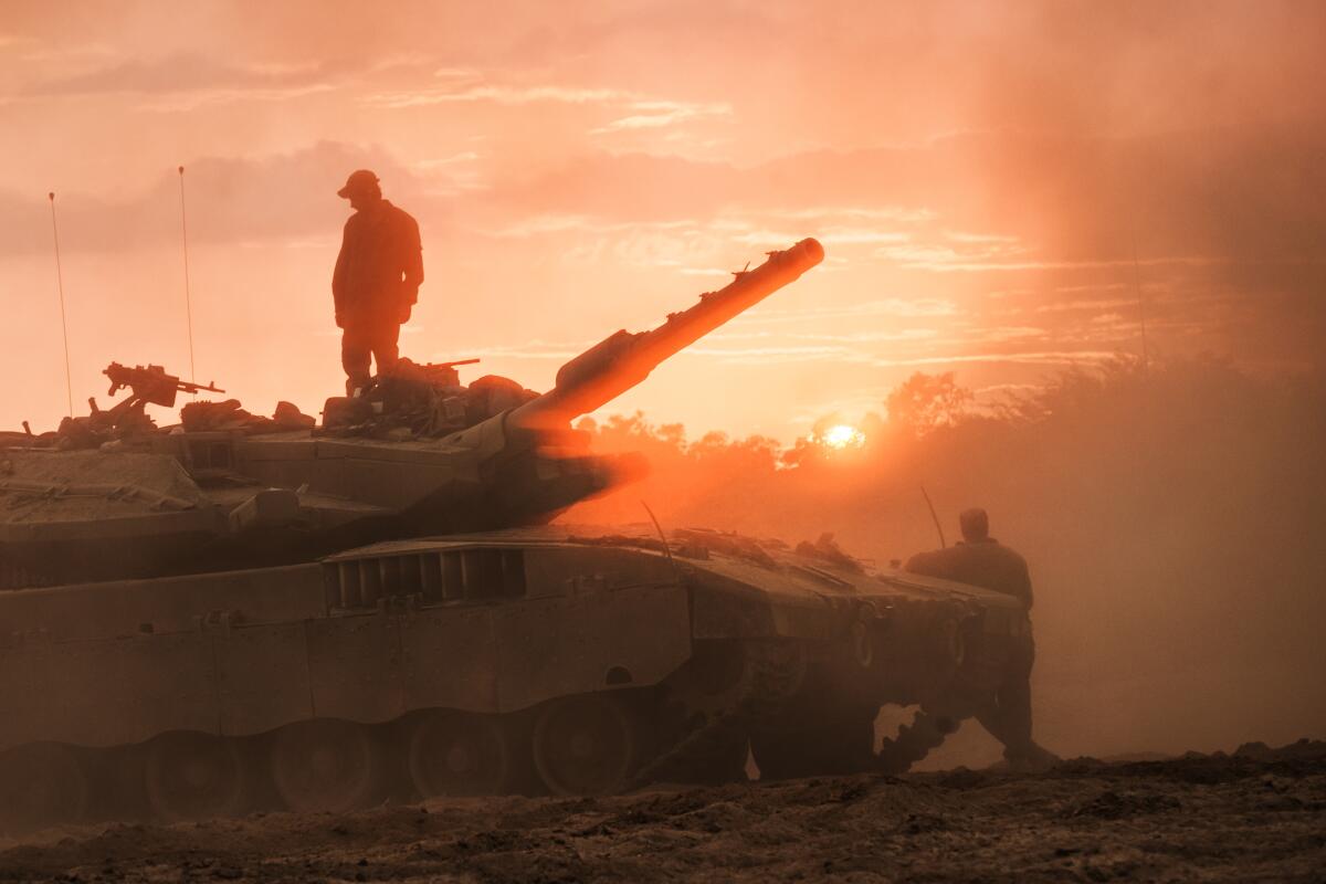 Soldiers stand on and near an Israeli battle tank near the border of Gaza, silhouetted against a hazy sun.