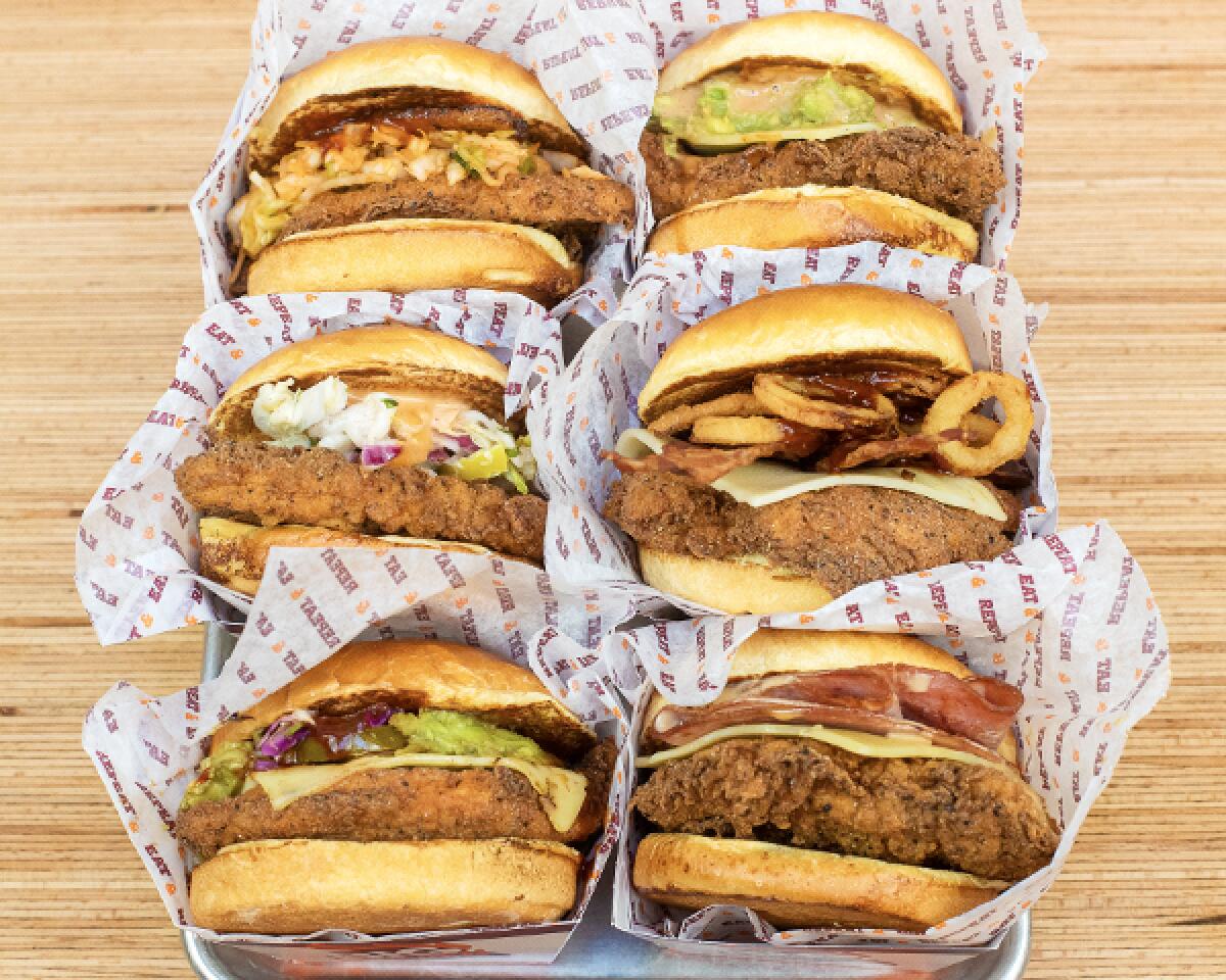 An overhead photo of a box of six fried chicken sandwiches all topped with varying accoutrements such as onion rings, cheese