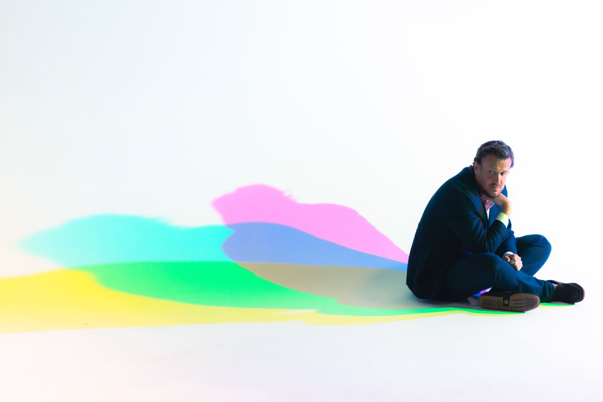 Jason Segel sits on a white floor with colorful shadows spreading out behind him.